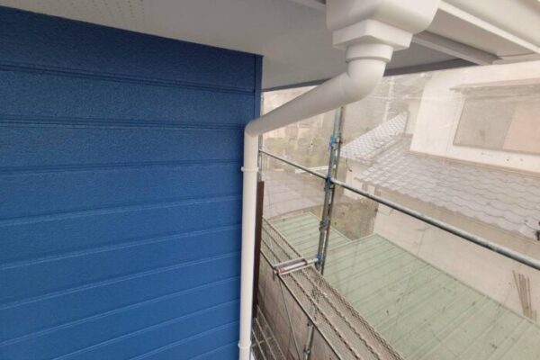 青く塗装された外壁と新しく交換された白い雨樋。軒樋から縦樋まできれいに接続され、足場の上から撮影された外壁塗装と雨樋交換後の様子。