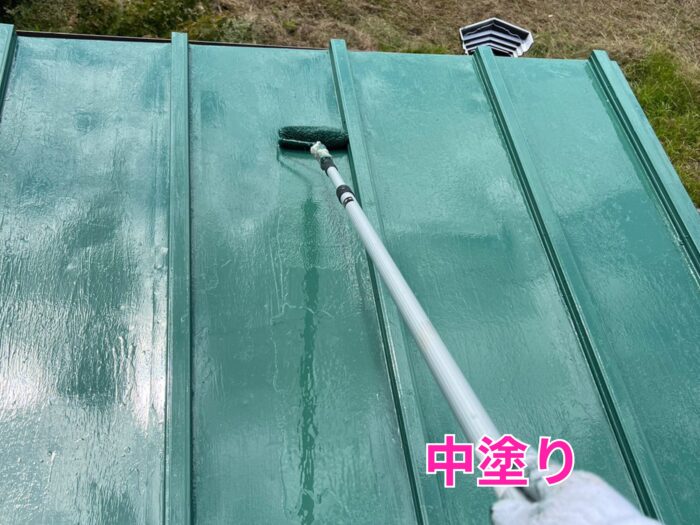 緑色の金属屋根をローラーで均一に塗装している中塗り工程の写真。塗膜が光沢を帯びている。