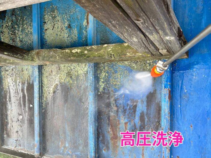 金属屋根の表面に付着した苔や汚れを、高圧洗浄機のノズルから勢いよく噴射される水で落としている様子。青い屋根材の上に苔が広がり、洗浄で表面がきれいになっていく途中の状態。